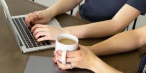 person holding a mug sitting next to a person typing on a laptop.